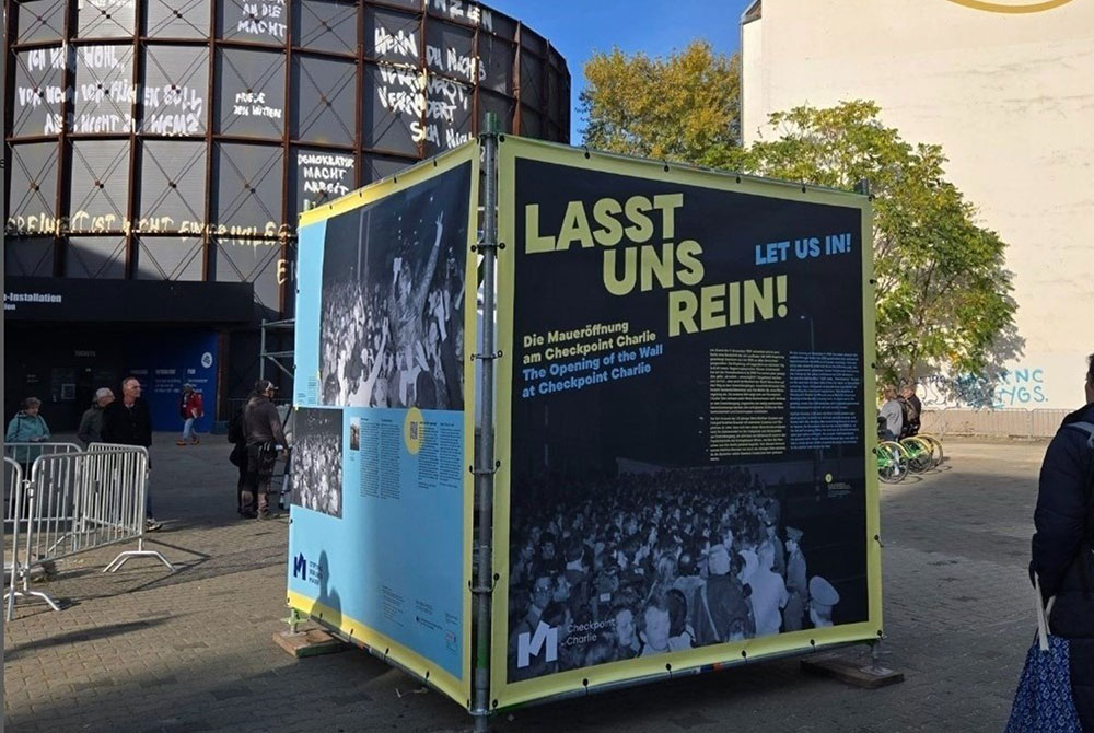 Bei gutem Wetter sind gelb-blaue Plakate zur Open-Air-Ausstellung „Lasst uns Rein!“ am Checkpoint Charlie zu sehen. Im Hintergrund ist das asisi-Panorama „Die Mauer“ zu erkennen. 